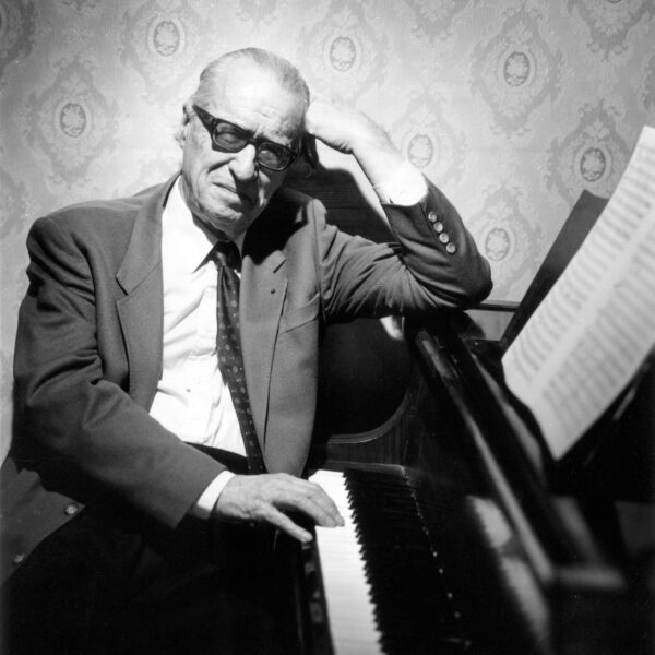 Composer Jean Françaix seated at the piano, leaning on the music stand with one arm, the other hand resting on the keyboard